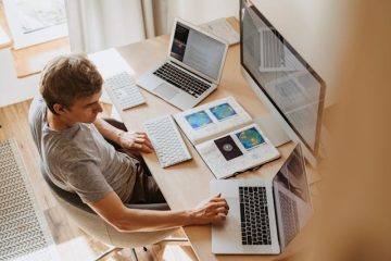 A young man engaged in hybrid work at a desk, using multiple electronic devices including two laptops, a desktop monitor, and an open notebook displaying data visualisations. He is focused on his tasks, working with a wireless keyboard and mouse in a well-lit home office setting